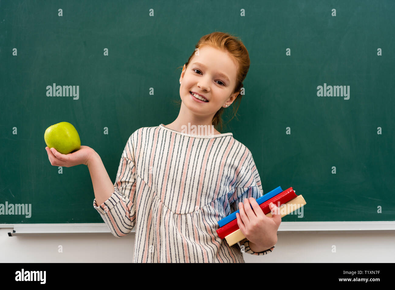 Cheerful ginger shoolgirl holding books and green apple in front of ...