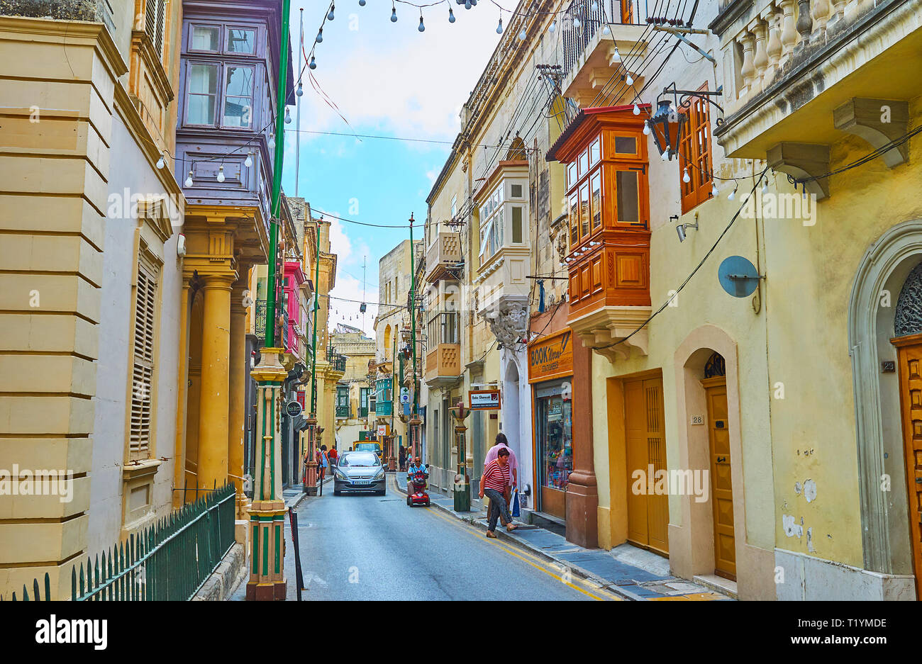 RABAT, MALTA - JUNE 16, 2018: The urban scene in old town with driving car, walking pedestrians, small stores, city festival decorations and lamps, on Stock Photo
