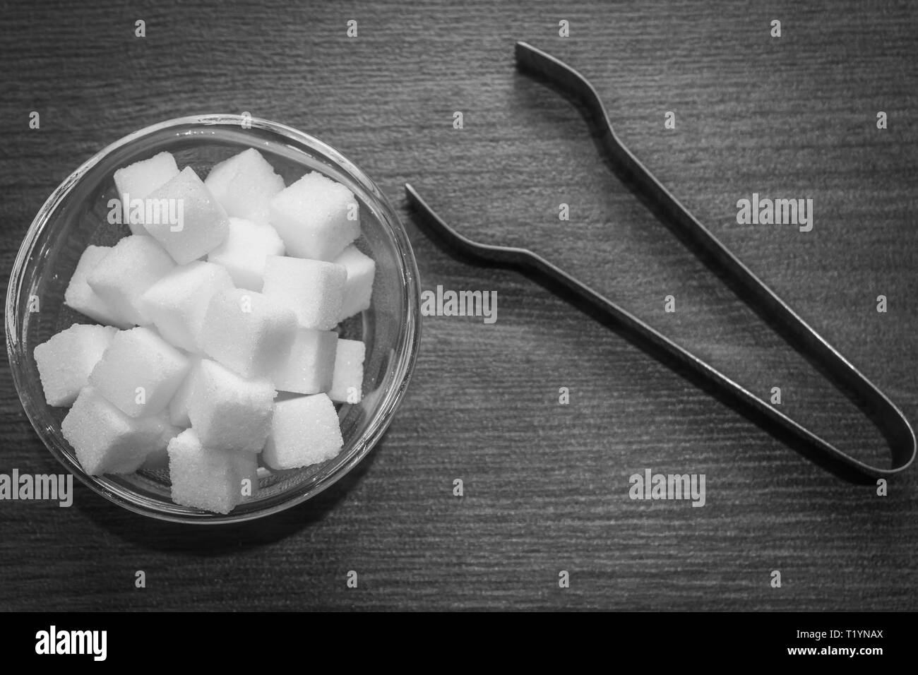 Top view of sugar cubes in a glass bowl with a pair of kitchen tongs foreceps kept on wooden floor. Contrasty black and white kitchen setup. Stock Photo