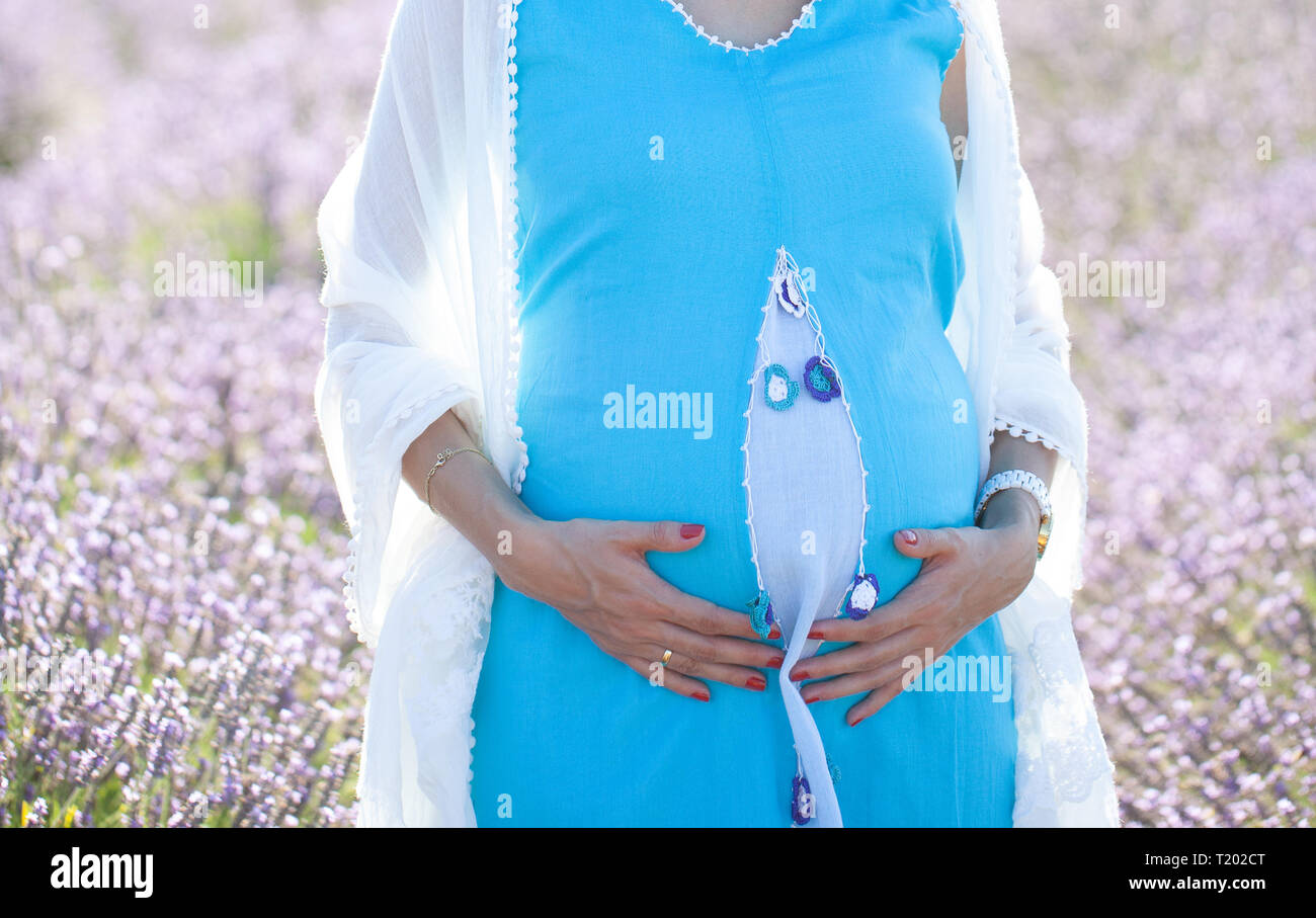 Cropped image of pregnant woman in blue traditional turkish dress and hugging the tummy in lavender field. Stock Photo