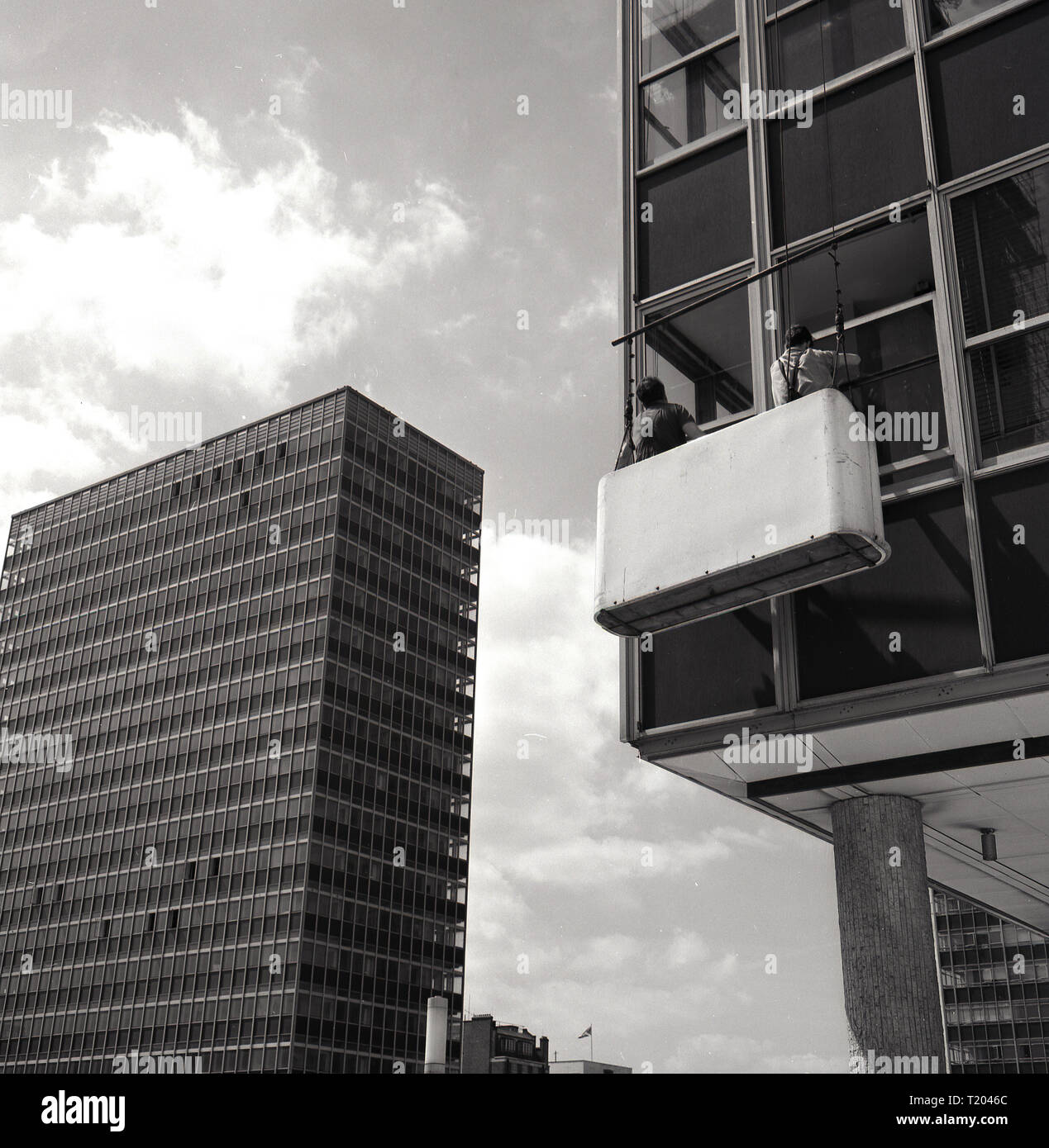 1960s, historical, two male window cleaners standing in a pod or vessel suspended from the roof, cleaning the windows of a modern high rise office building, London, England, UK. Stock Photo