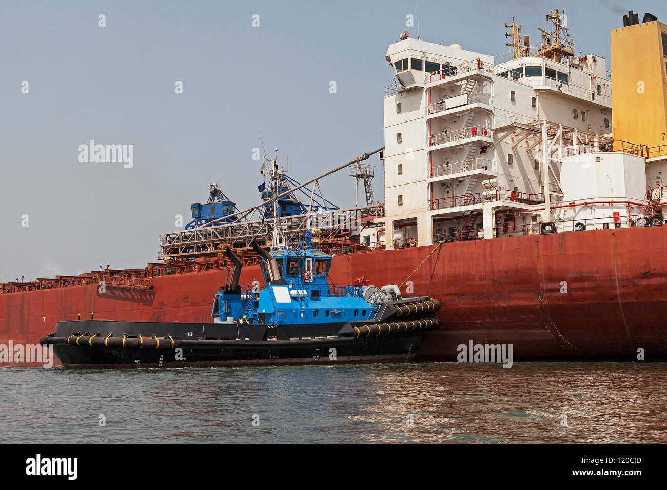 Port operations for managing and transporting iron ore.Tug manoeuvring and turning transhipment vessel before docking and loading up ore at jetty Stock Photo