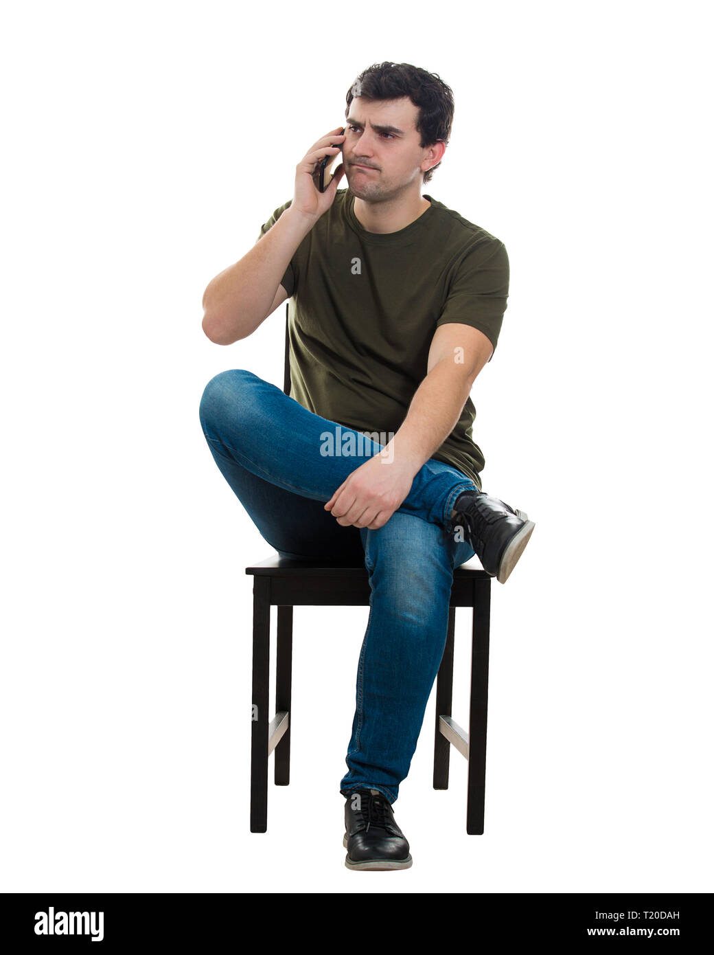 Full length portrait of frustrated young man sitting on chair talking on his mobile phone. Unhappy casual guy receiving bad news on the telephone isol Stock Photo