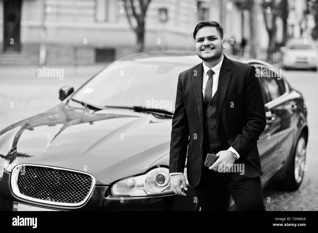 Stylish indian businessman in formal wear with mobile phone standing against black business car on street of city. Stock Photo