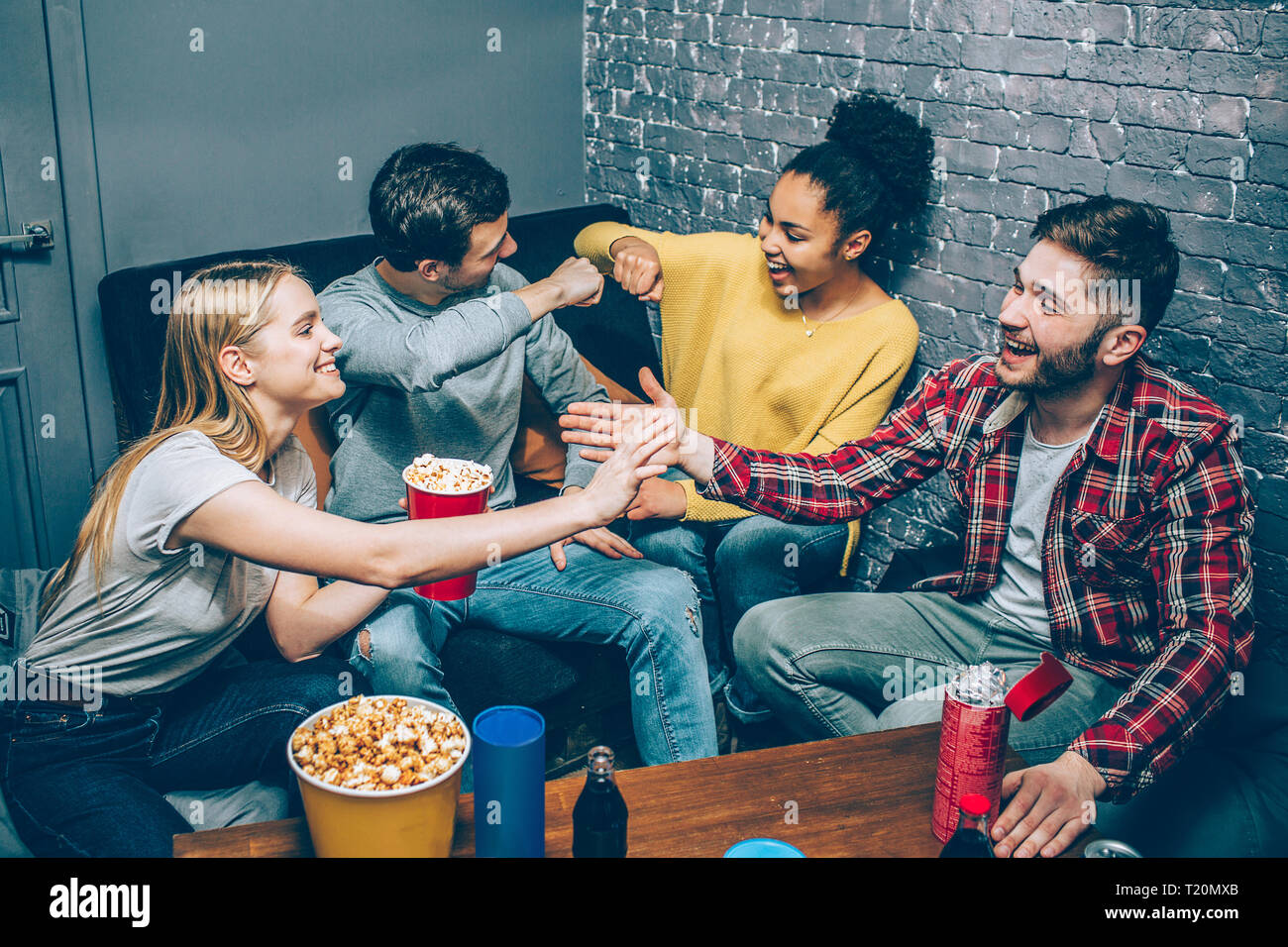 A picture of young boys and girls sitting together in a small room on couches and greeting each other. They got some popcorn and ready to play some gr Stock Photo