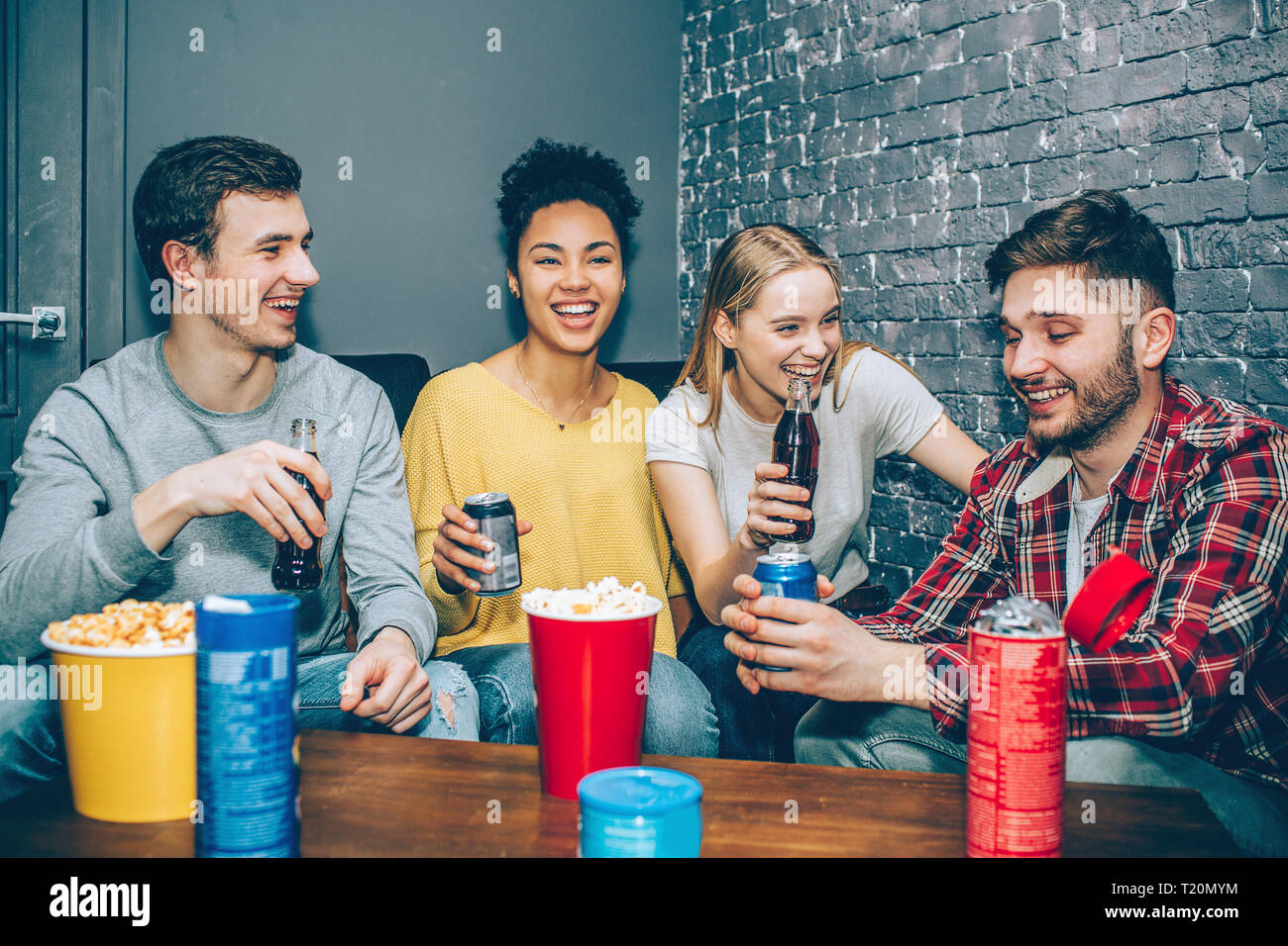 Energetic and happy young people are sitting in small grey room eating popcorn and drinking some cola. They are ejoying time spending together Party n Stock Photo