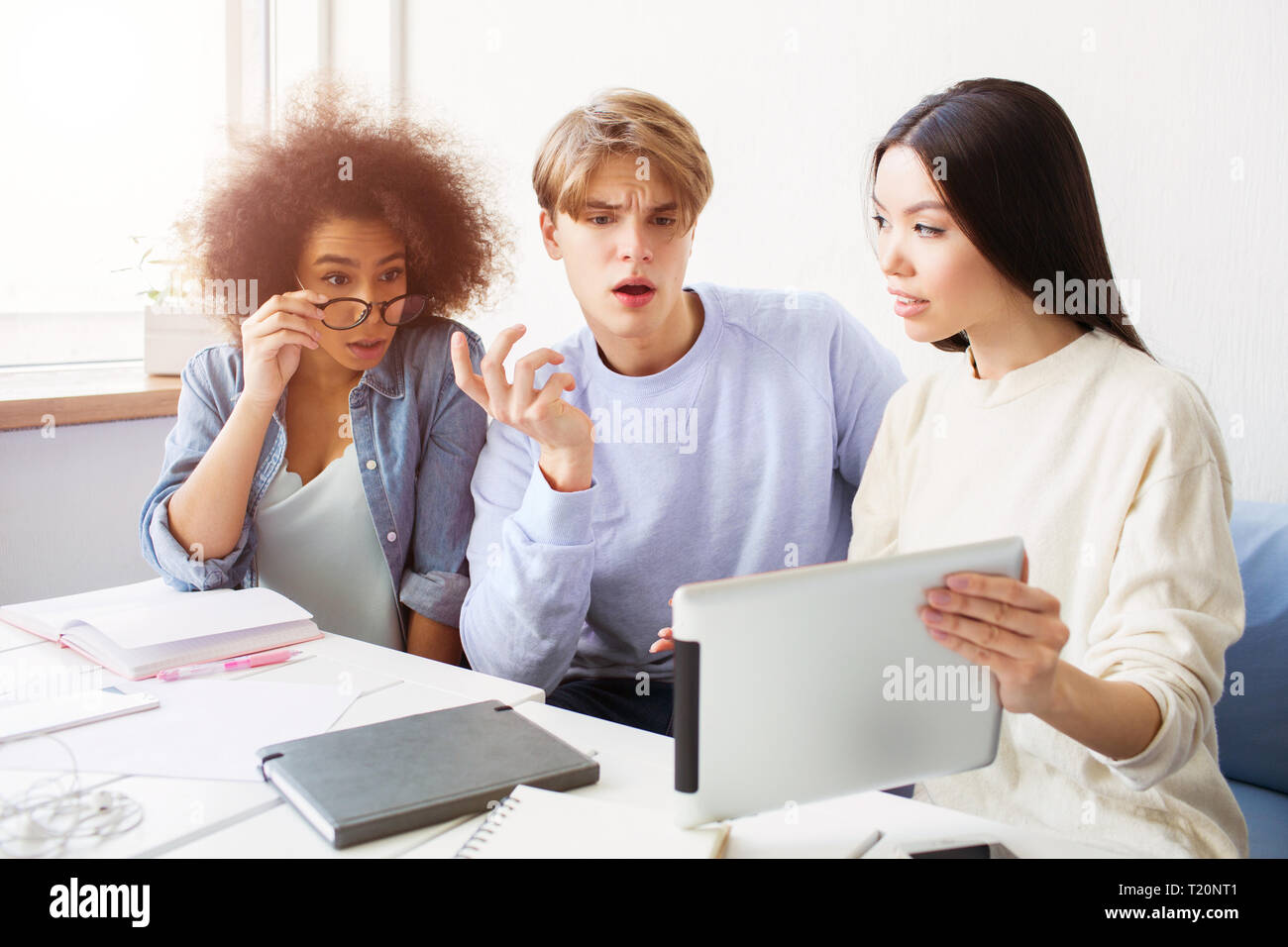 Three wondering people are sitting at the table and studying. They are amazed. They can't believe in what they see on screen. Asian girl is holding a  Stock Photo
