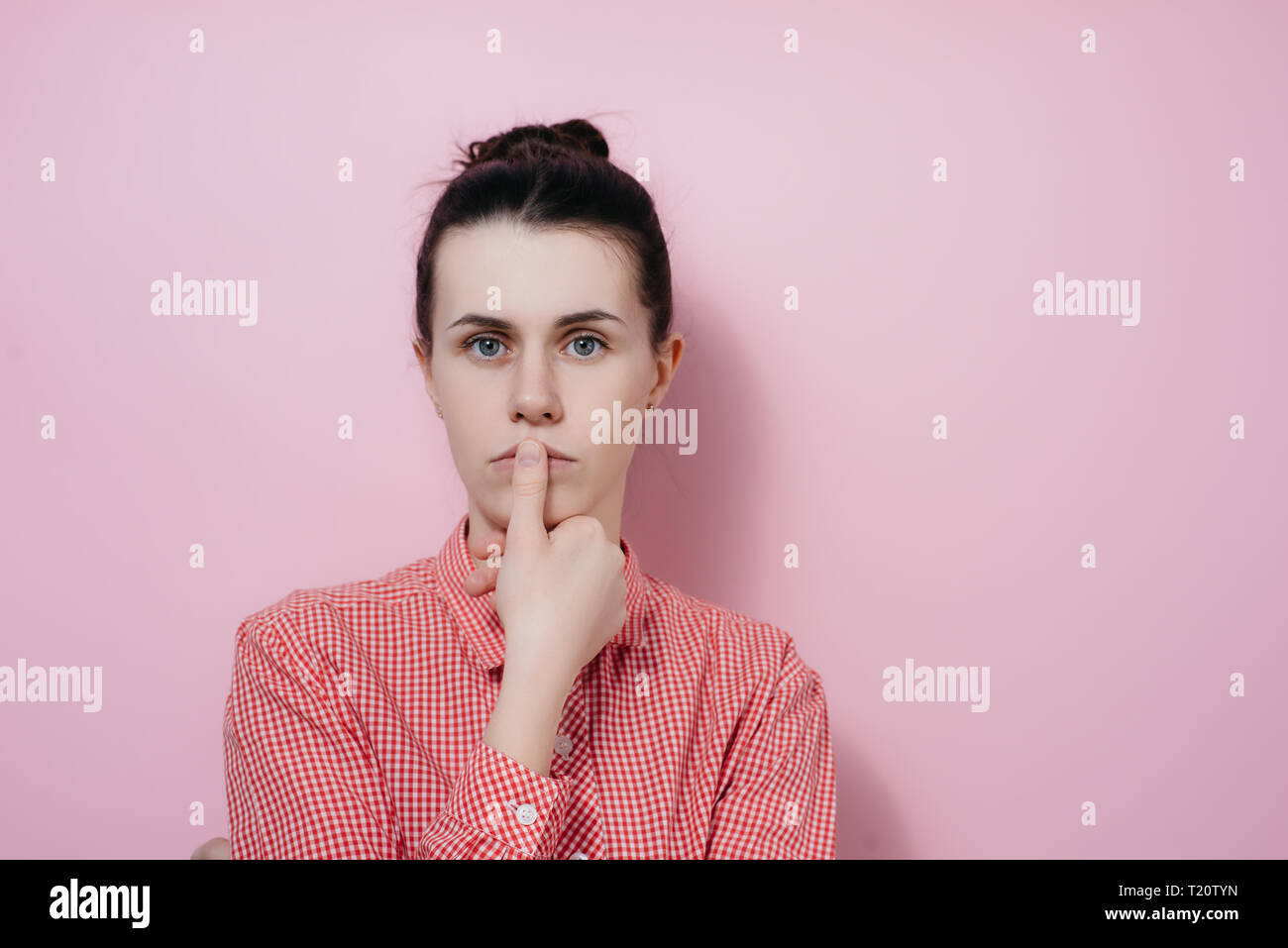 Close up studio portrait beautiful female with puzzled look, keeps finger near lips, looks at camera, has shocked expression, dressed in shirt, posing Stock Photo