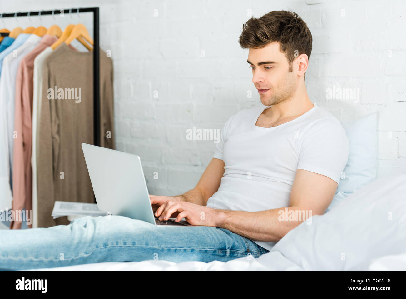 man in white t-shirt and jeans sitting on bed and typing on laptop in bedroom Stock Photo
