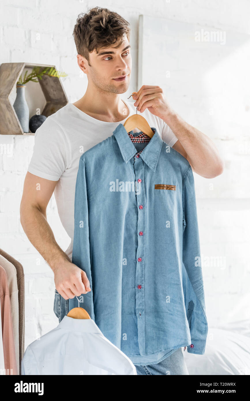 handsome man in white t-shirt holding and trying on shirts in bedroom Stock Photo