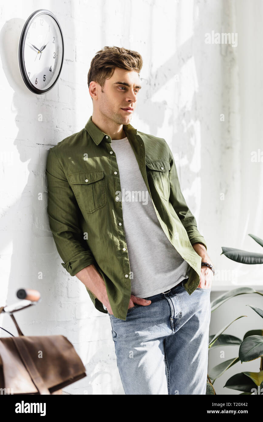 handsome man in green shirt and jeans standing near brick wall, clock and  plants in office Stock Photo - Alamy