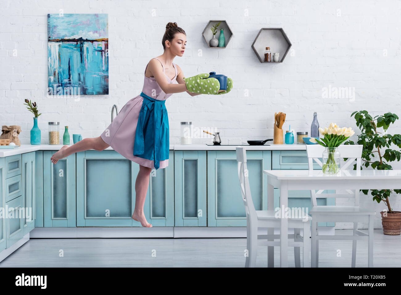 beautiful barefoot girl in potholders levitating in air with pot in kitchen Stock Photo