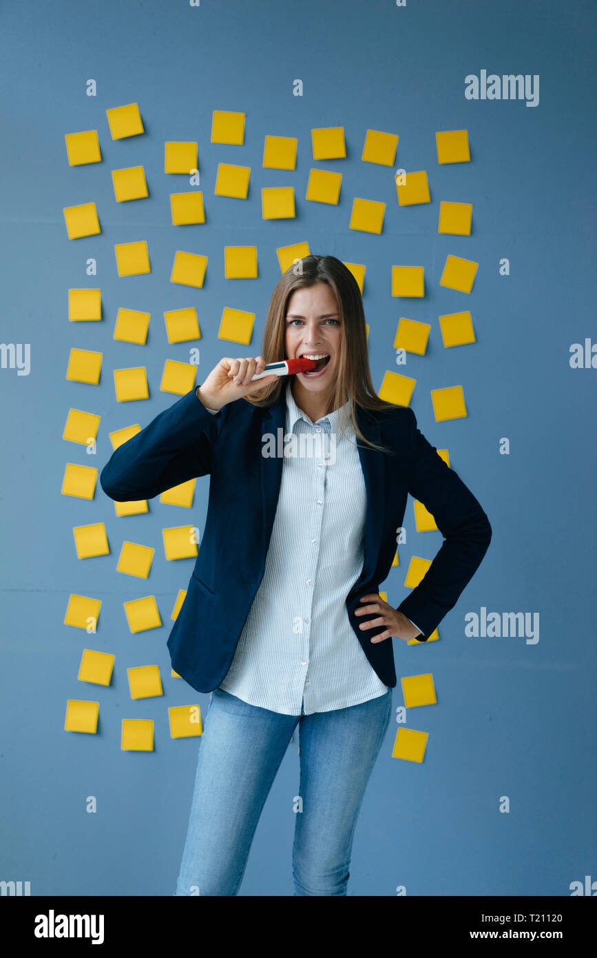 Yong businesswoman standing in front of wall, full of yellow sticky notes, biting marker pen Stock Photo
