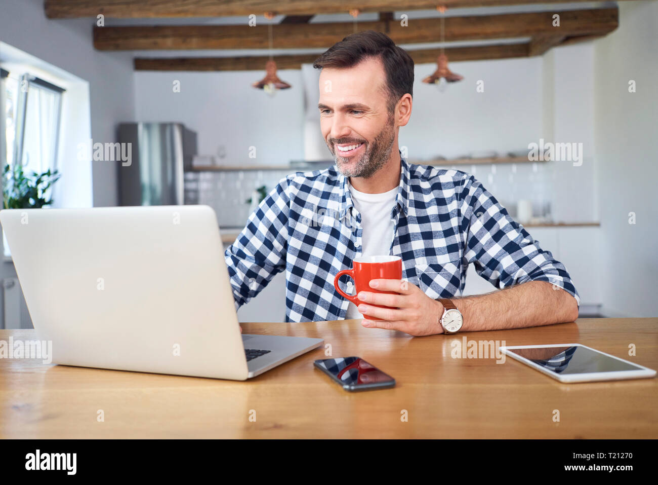 Smiling man working remotely on laptop at home and having coffee Stock Photo