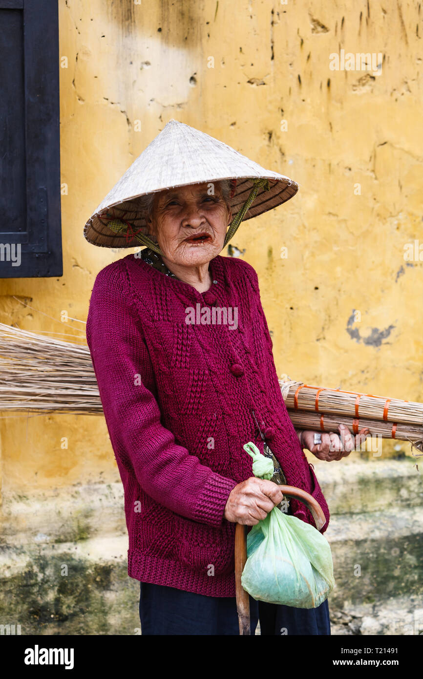 Old woman wearing conical hat and carrying reed brooms  Vietnamese hat pauses to pose for photographer while shopping Stock Photo