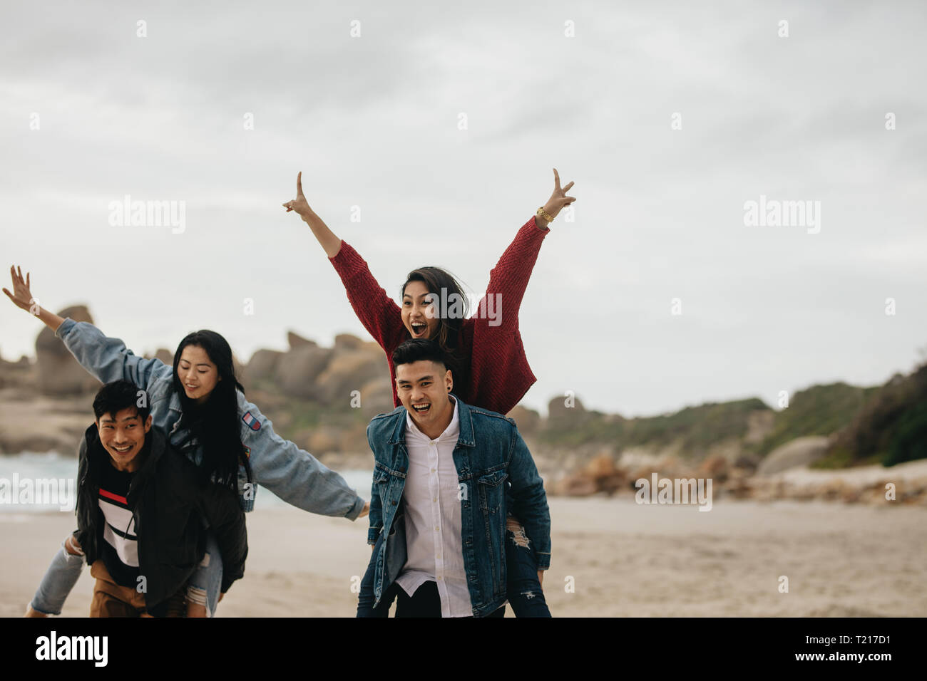 Two young couples walking by the beach, with men carrying their women on their back. Couples piggybacking on sea shore. Stock Photo