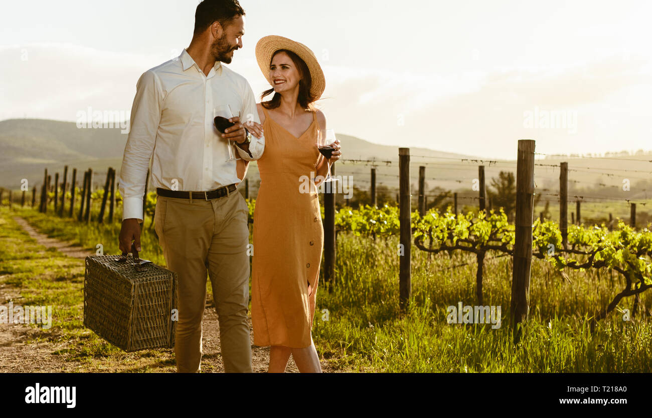 Smiling couple walking in a wine farm drinking red wine and carrying a picnic basket. Couple in love on a date walking in a vineyard holding each othe Stock Photo