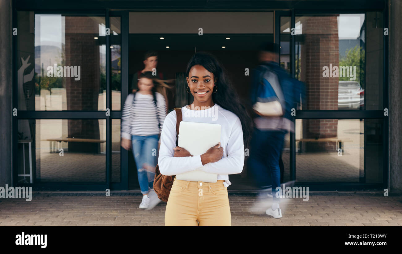 Portrait of girl student standing at university campus with other students walking in background. Young woman standing in college with students walkin Stock Photo