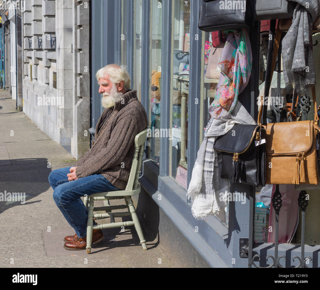 grey haired man sitting on a chair by a shop window. Stock Photo