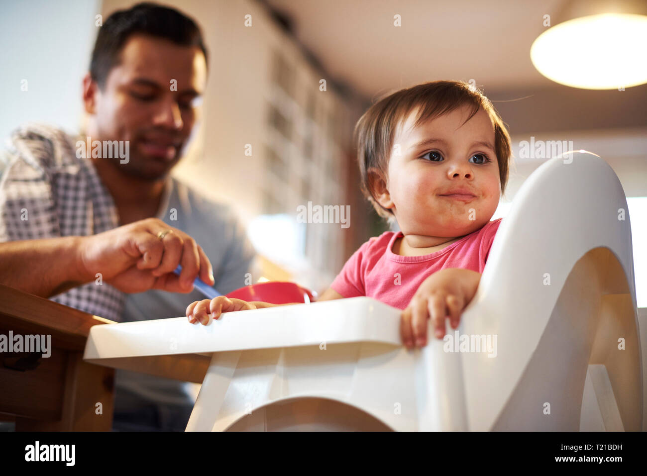 Portrait of baby girl sitting in high chair at home with father in background Stock Photo Alamy