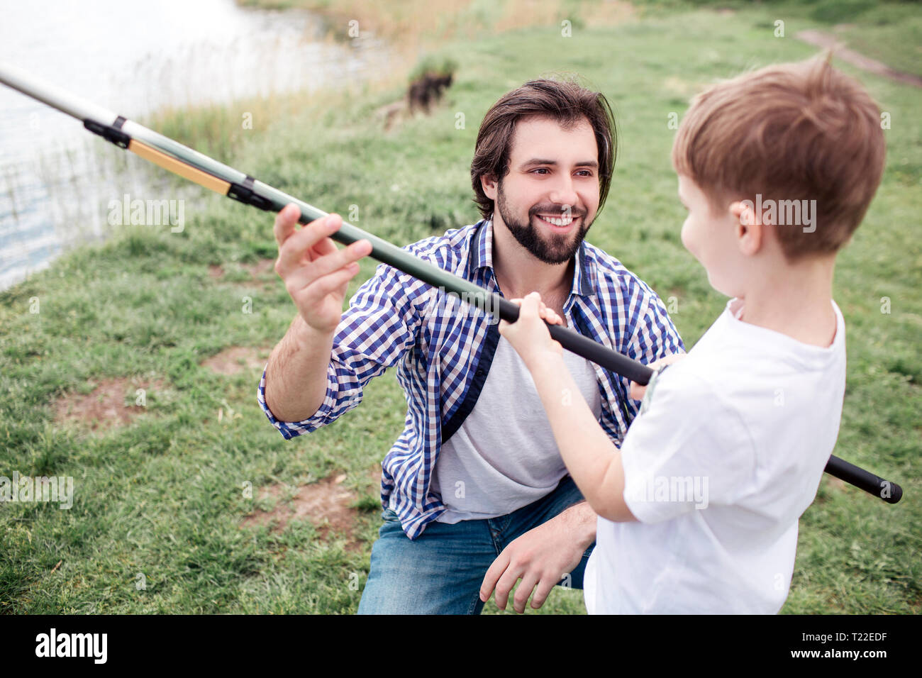 Happy dad is giving fish-rod to his son. He is smiling. Boy is holding fish-rod very tight and looking at dad Stock Photo