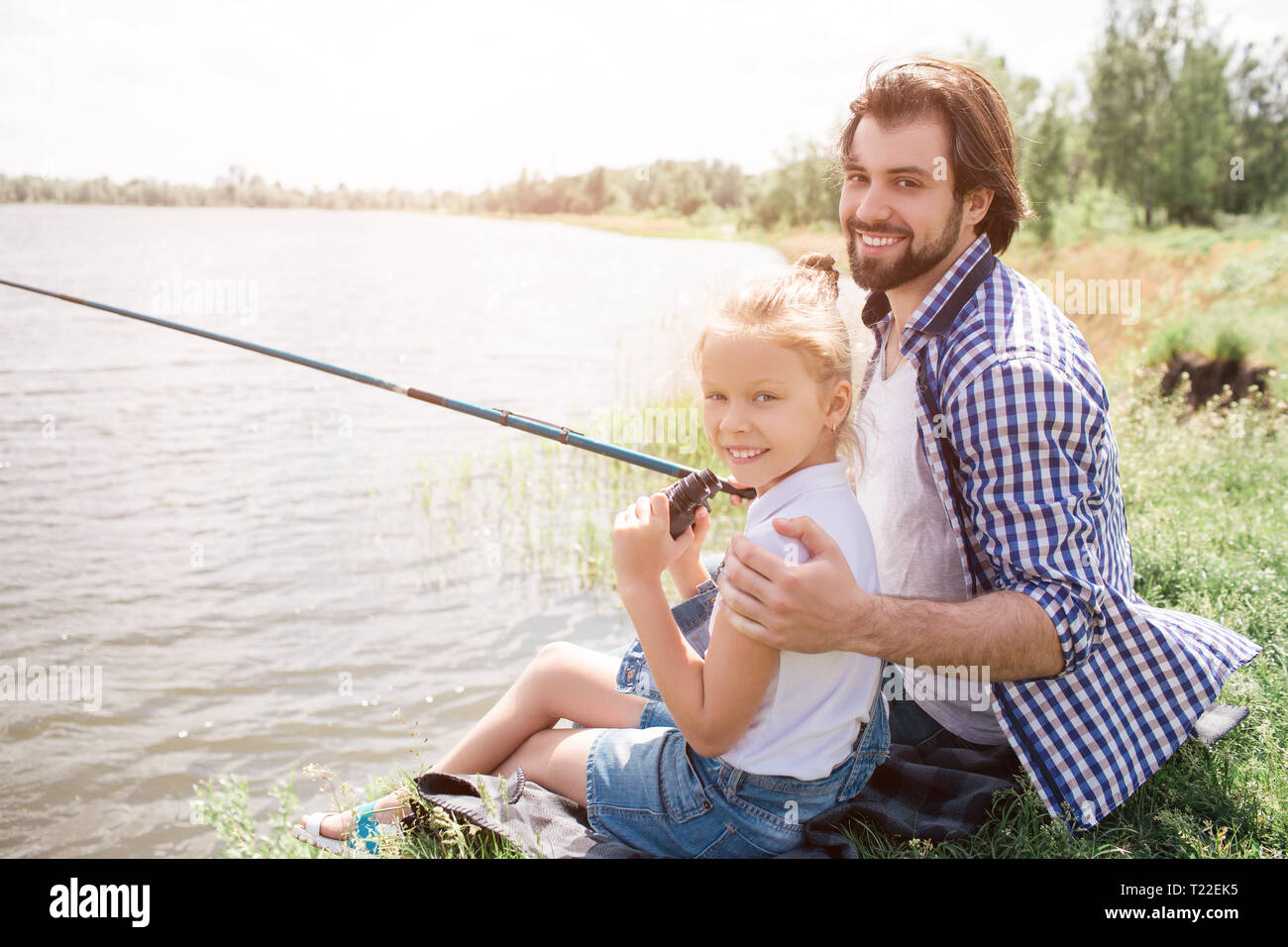 Happy dad and daughter are sitting on grass near water and looking at  camera. He is huging her and holding fish-rod in right hand. They are  fishing Stock Photo - Alamy