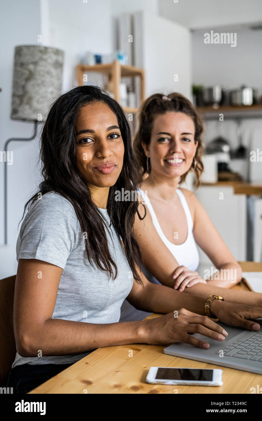 Portrait of two smiling women sitting with laptop at table at home Stock Photo