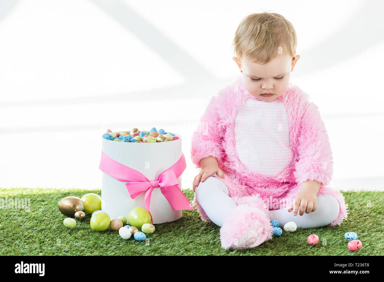 cute baby in pink fluffy costume sitting near box with colorful Easter ...