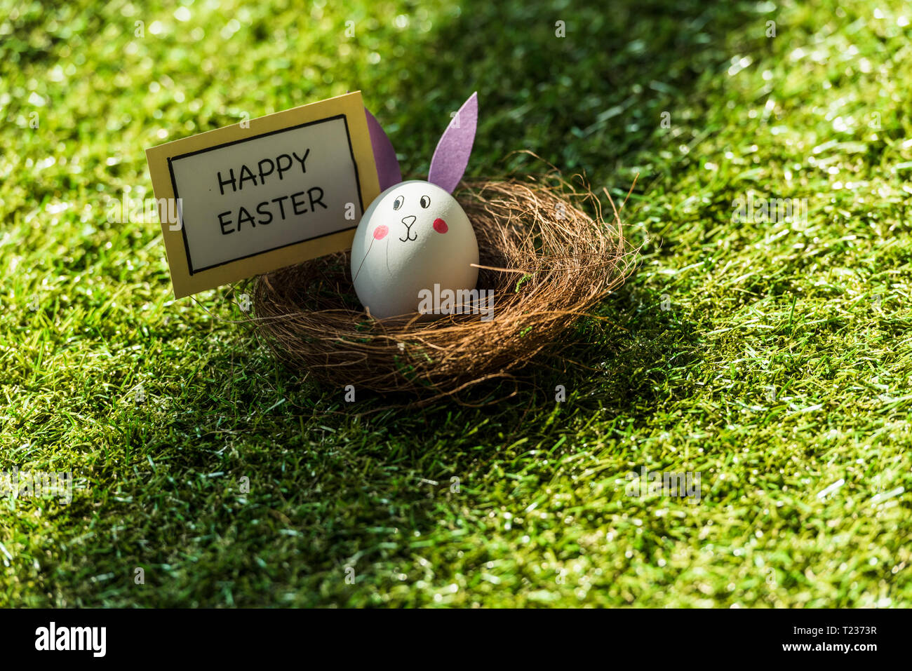 chicken egg with drawn rabbit face and paper ears, and happy Easter card on green grass Stock Photo