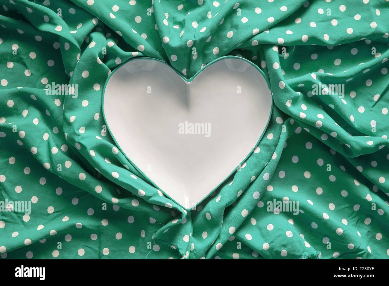 Stoneware heart shaped plate with emerald green edge on green and white polka dot printed fabric fun romantic background texture. Stock Photo