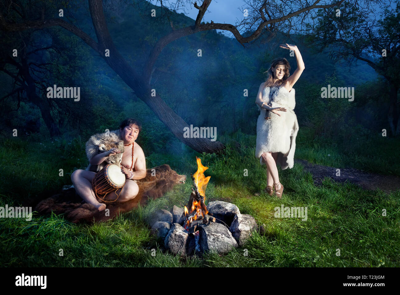 Caveman dressed in wolf skin playing drum and cave woman dancing near bonfire in the forest Stock Photo