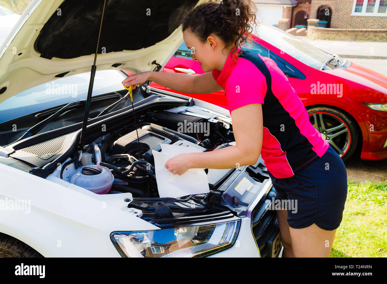 Young Caucasian female in her 20’s under the bonnet checking the oil level on her car using the dipstick and a cloth. Stock Photo