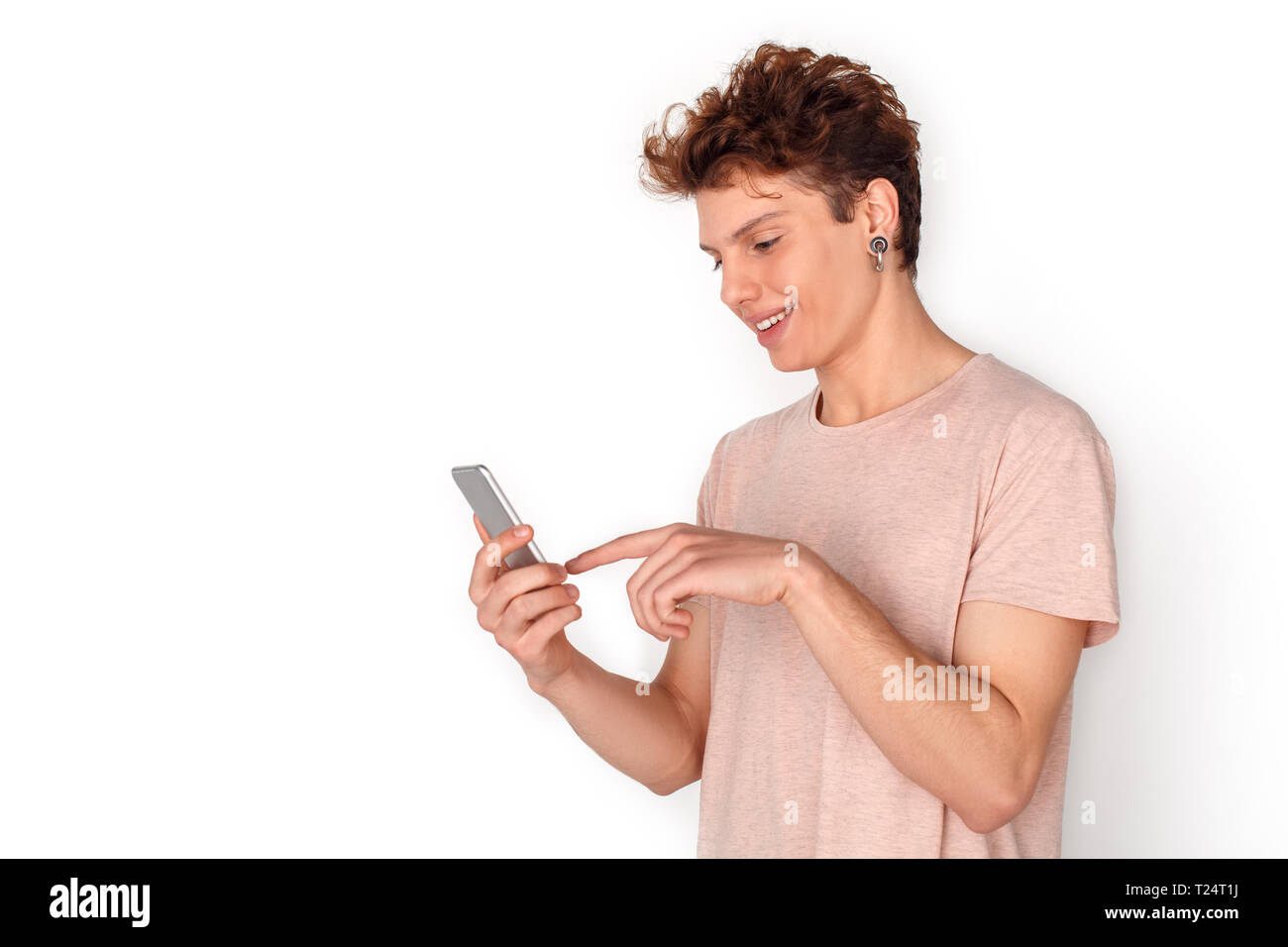 Teenager boy wearing earrings studio standing isolated on white wall browsing internet on smartphone smiling joyful side view Stock Photo