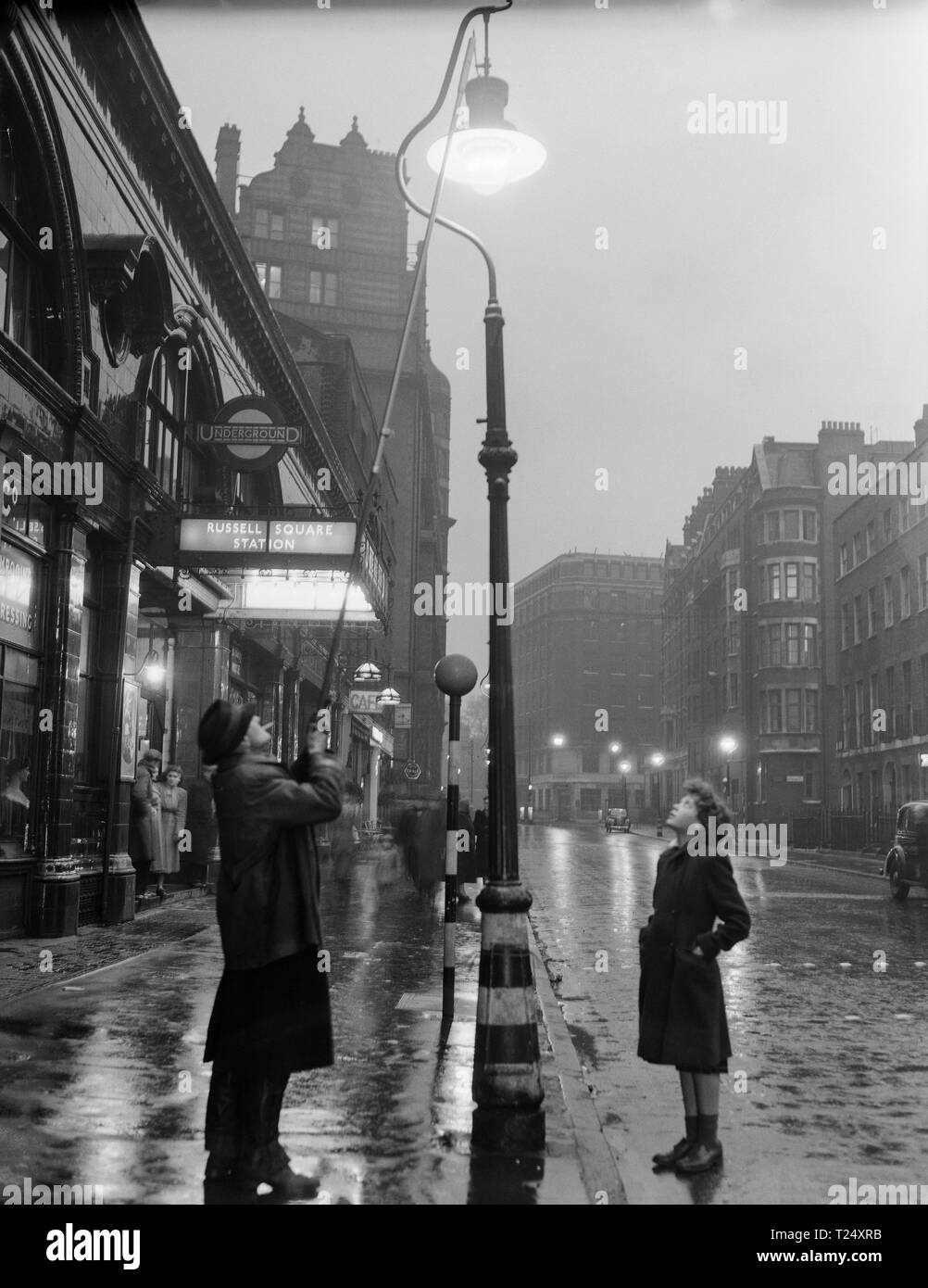 Black and white photograph taken in 1950 outside Russell Square Underground Tube station in London, England. It shows a man lighting a gas street lamp, watched by a young girl. Stock Photo