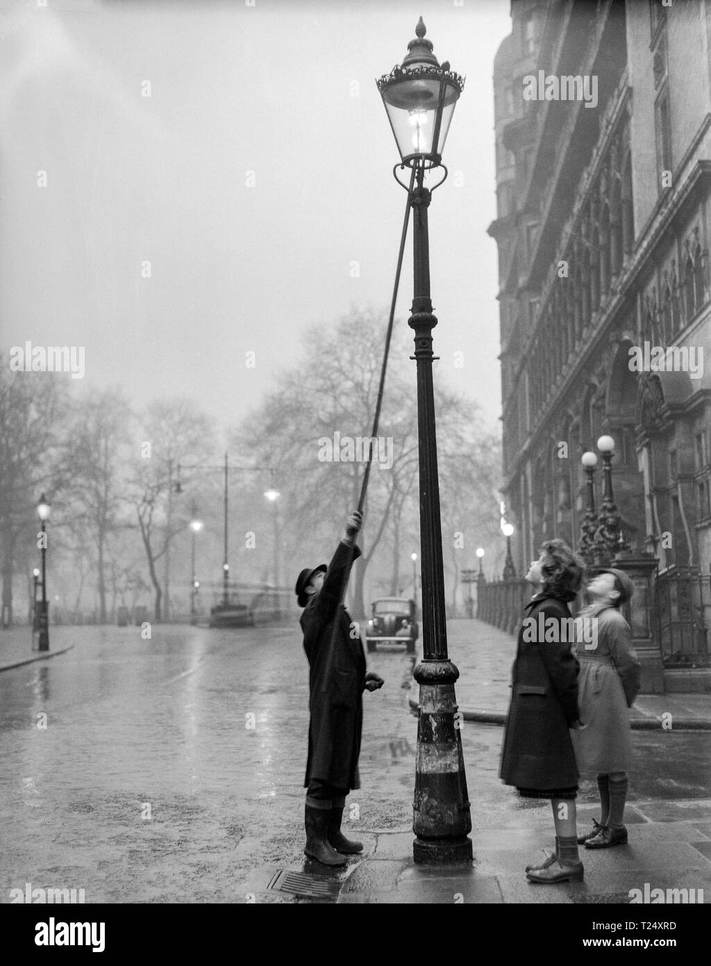 Black and white photograph taken in 1950,  in London, England. It shows a man lighting a gas street lamp, watched by a young girl. and young boy. Stock Photo