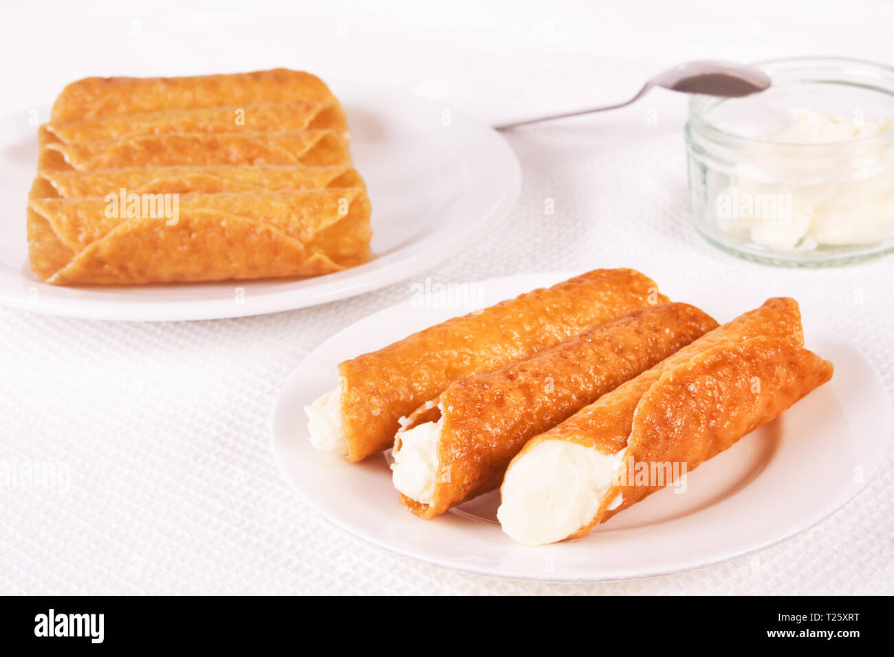Cream filled brandy snaps served on a plate. Unfilled snaps and a pot with cream in the background Stock Photo