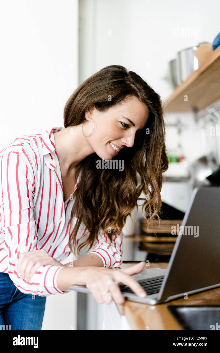 Smiling woman using laptop in kitchen at home Stock Photo