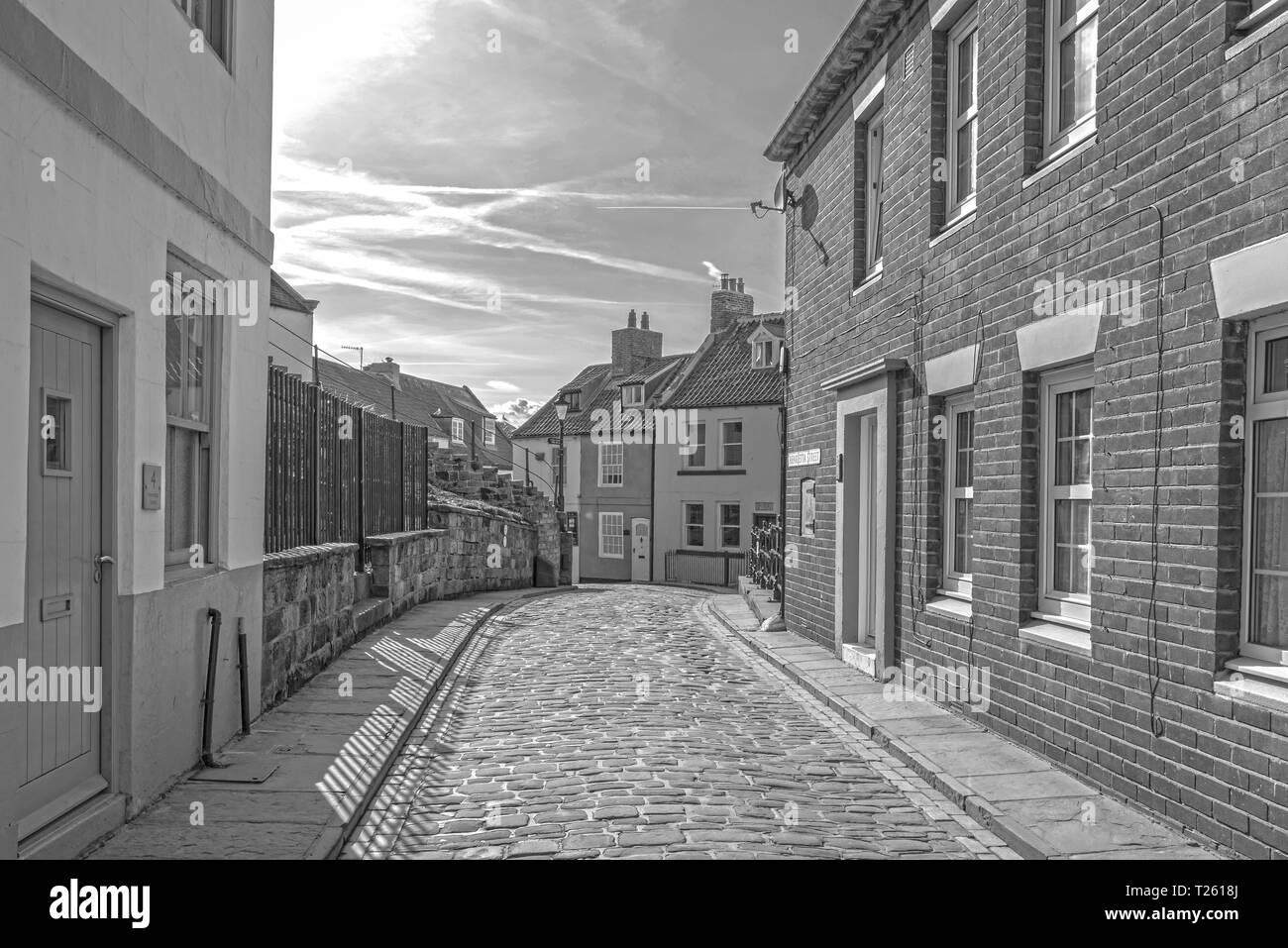 Lower end of Henrietta Street in Whitby.  Houses are on each side of a cobbled street with a lamppost at the end. A blue sky is above. Stock Photo