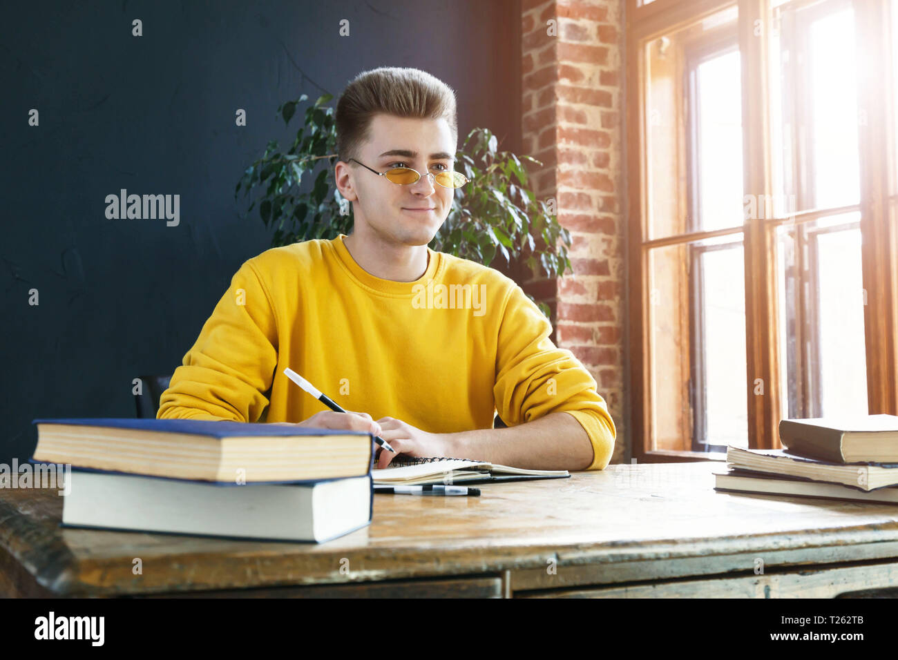 Young serious blonde man wearing glasses and yellow pullover studying or working in the class Stock Photo
