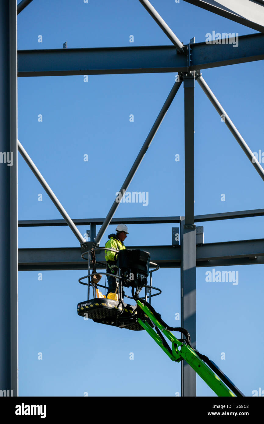 Steel frame of new building in construction with construction worker on crane lift inspecting joint set against bright blue sky. Stock Photo