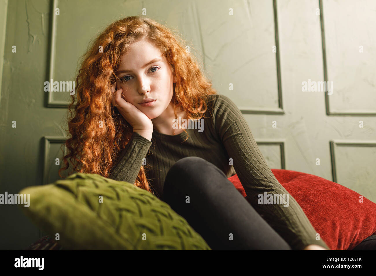 Curly redhead girl propped up her head and sitting at the sofa around the pillows in the green interior Stock Photo