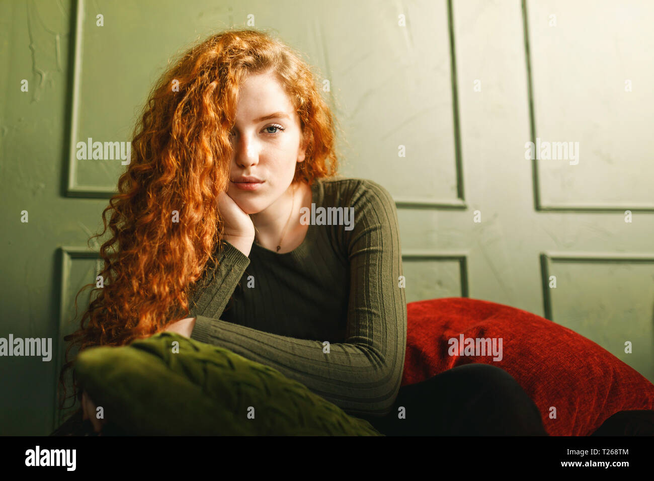 Curly ginger girl propped up her head and looking to the camera while sitting at the sofa around the pillows in the green interior Stock Photo