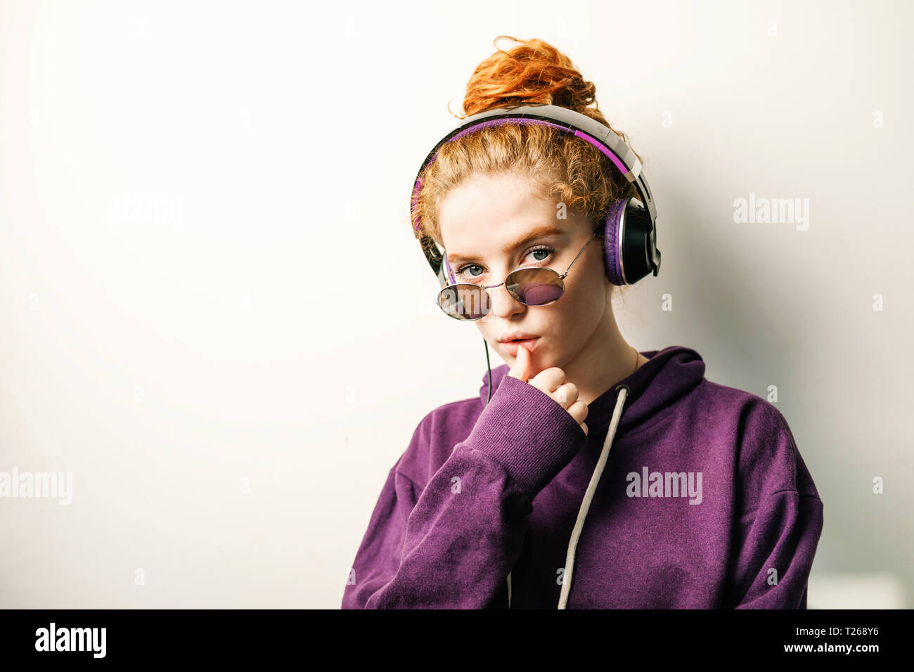 Curly ginger girl in headphones wearing glasses and purple hoodie listening her favorite music Stock Photo
