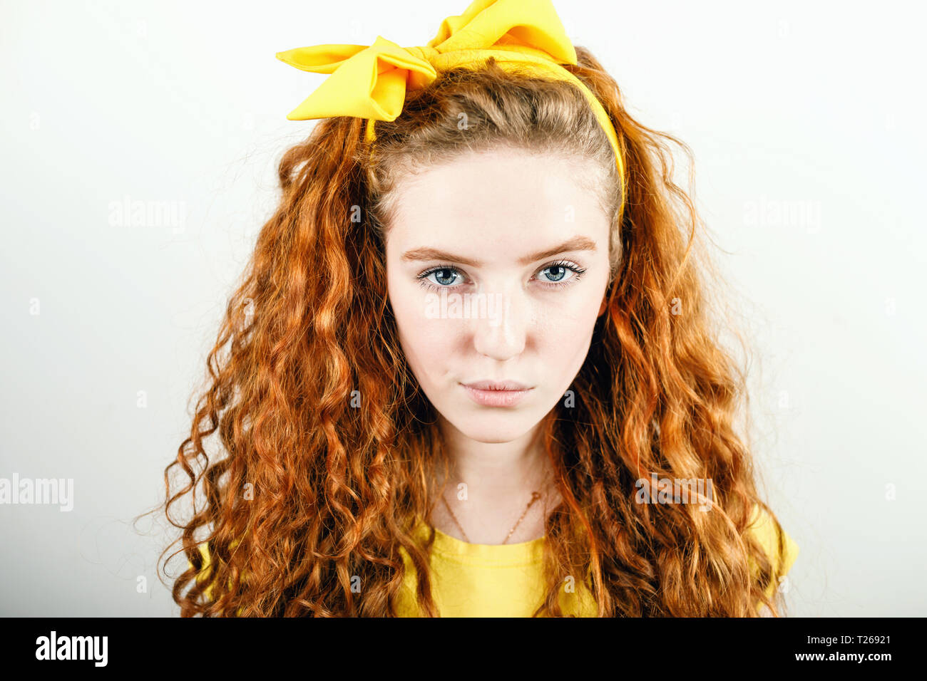 Portrait of curly redhead girl with a yellow bow on her head wearing yellow t-shirt on the white background Stock Photo