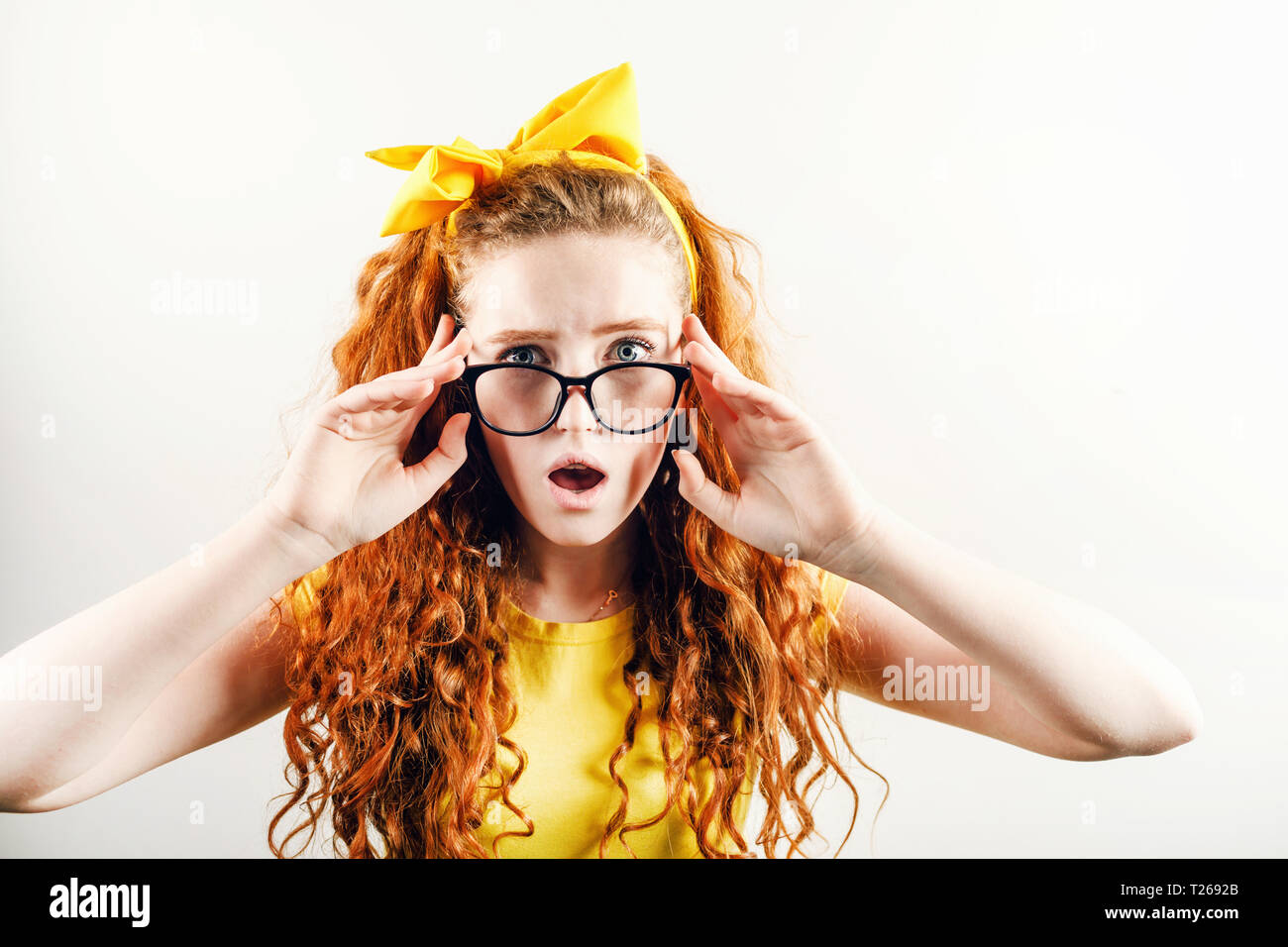 Surprised curly redhead girl in glasses with a yellow bow on her head wearing yellow t-shirt looking to the camera with schoked emotion Stock Photo