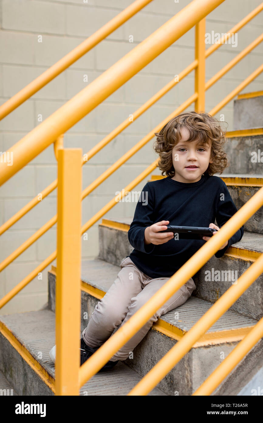 Portrait of boy sitting on stairs with handheld game console Stock Photo