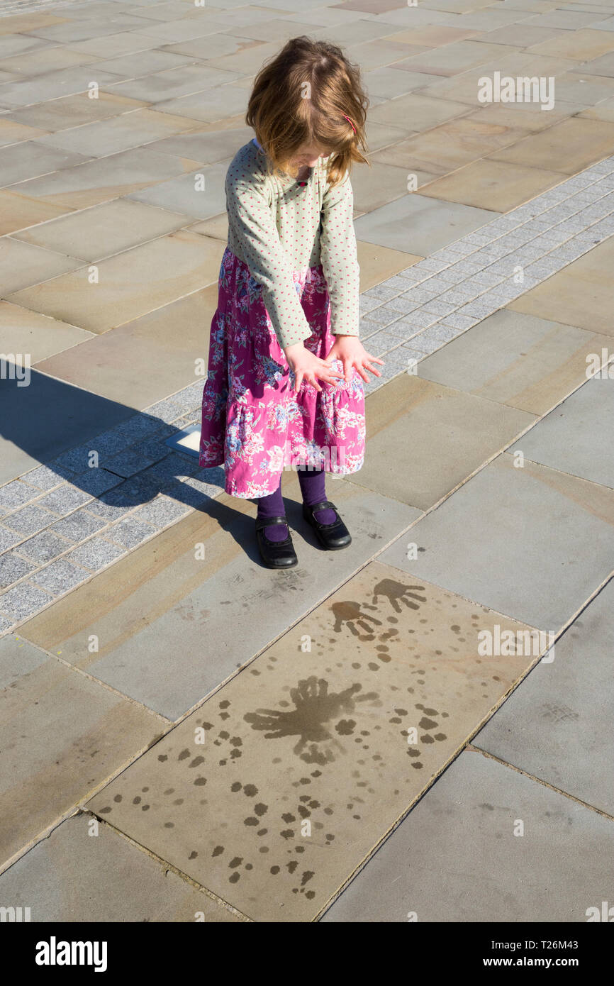 Four-year-old girl makes handprints with water from the water feature inside the quadrangle of The Piece Hall. Sun sunny day. Halifax, West Yorkshire, UK. (106) Stock Photo