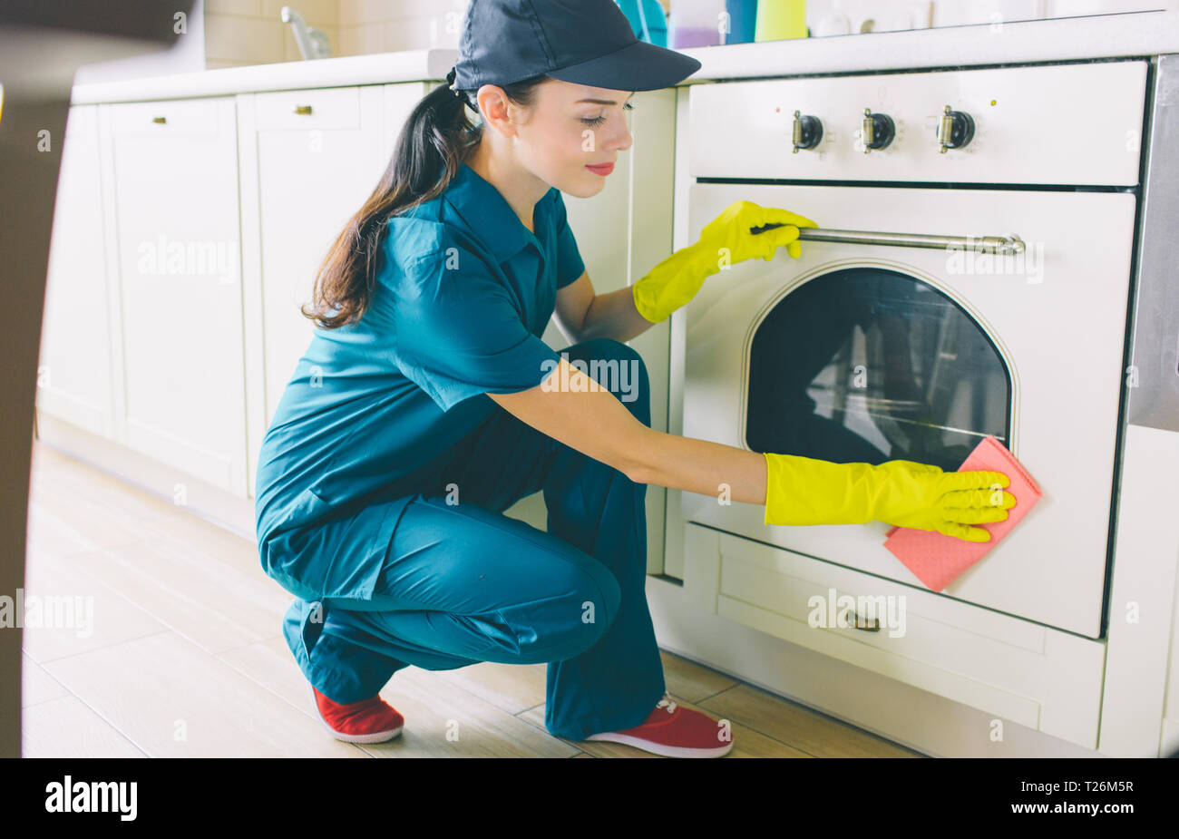 A picture of cleaner sitting in squad position and washing stove. She uses orange rag. Girl cleans very good. She is a hardworker Stock Photo