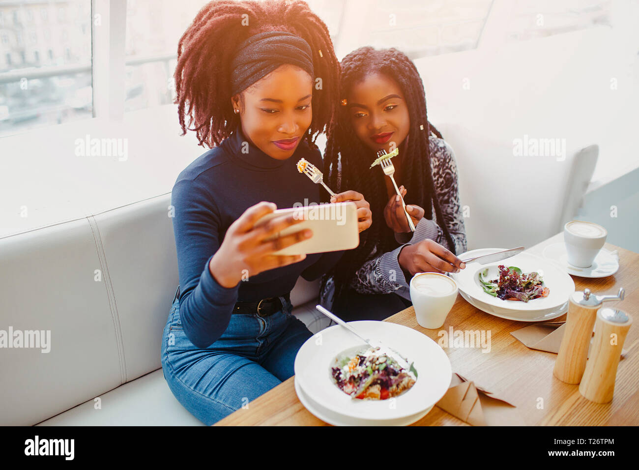 Two african models sit and pose on phone camera. They hold forks with food close to mouth. People are in cafe Stock Photo