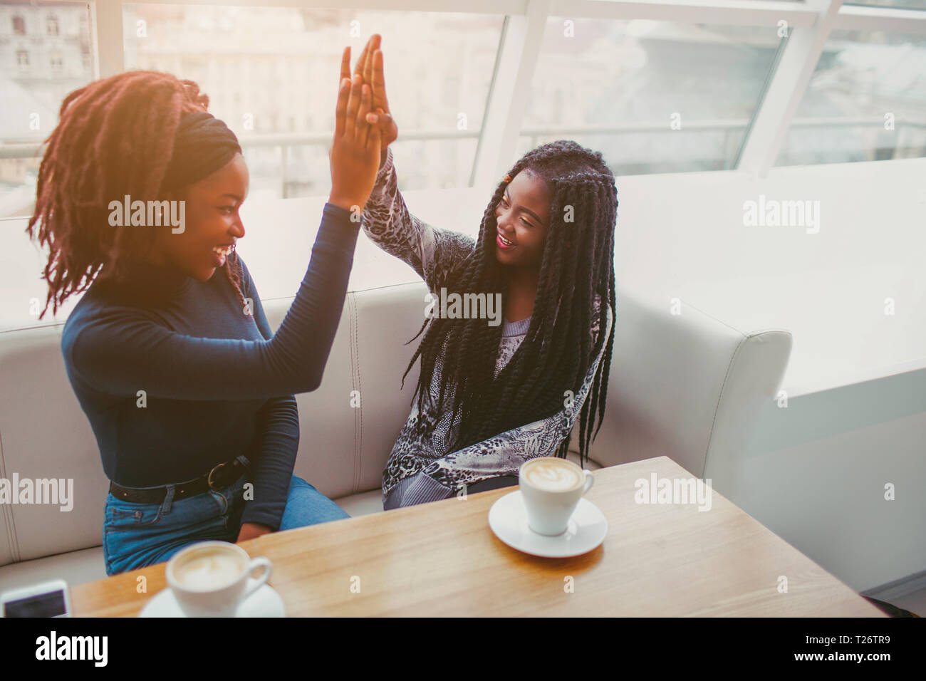 Tow african models give high five one to another. They look at each other and smile. Frieds spend time in cafe at window Stock Photo